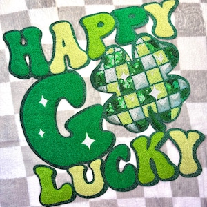 May include: A St. Patrick's Day design featuring the words "HAPPY" and "LUCKY" in green and yellow, with a sequined shamrock. The design is set against a checkered gray and white background.