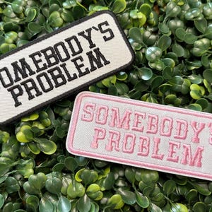 May include: Two rectangular embroidered patches with the text "SOMEBODY'S PROBLEM". One patch has black text on a white background with a black border. The other has pink text on a white background with a pink border. The patches are on a bed of green leaves.