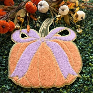 May include: Orange and purple pumpkin-shaped decorative item with a gold trim. The pumpkin has a textured surface and a large purple bow. The background includes small pumpkins and greenery, suggesting a fall or harvest theme.