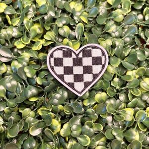 May include: A heart-shaped patch with a black and white checkered pattern. The patch has a white border and is set against a background of green foliage.
