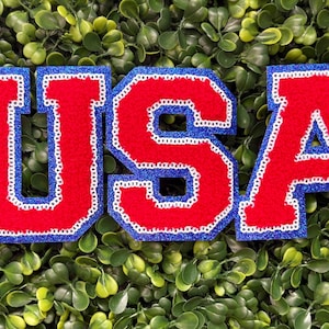 May include: A felt applique spelling "USA" in red letters with a white border and blue glitter outline. The letters are set against a background of green foliage.