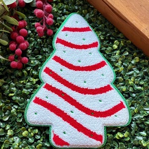 May include: A Christmas tree-shaped decorative item with a white background and red stripes. The tree has a green border and small green dots. The item is placed on a bed of green foliage.