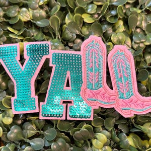 May include: Pink and teal sequined patches spelling "Y'ALL" with two pink cowboy boot patches. The patches are set against a backdrop of green foliage. The patches are likely for clothing or accessories.