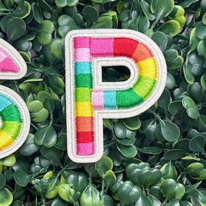 May include: Embroidered patches spelling out "SPF" in rainbow colors. Each letter is outlined in white and filled with a vibrant spectrum of hues, including red, pink, yellow, green, and blue. The patches are set against a backdrop of green foliage.