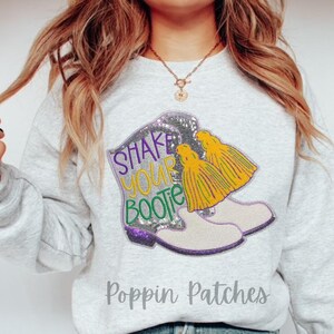 May include: A light gray sweatshirt with a colorful graphic of a pair of boots with the text "Shake Your Bootie". The boots have purple, silver, yellow, and green accents. The brand name "Poppin Patches" is printed at the bottom.