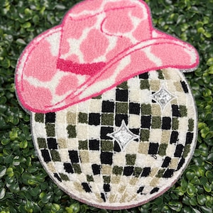 May include: A decorative rug featuring a pink and white cow print cowboy hat atop a disco ball design. The disco ball has a checkered pattern in black, white, and olive green, with two silver star accents.