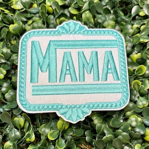 May include: Embroidered patch with the word "MAMA" in teal thread on a white background. The patch has a decorative teal border and is set against a backdrop of green foliage. A decorative accessory.