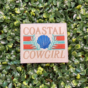 May include: Embroidered patch with the words "COASTAL COWGIRL" in tan thread. The design includes a blue seashell, red and teal stripes, and green leaf accents. The patch is rectangular and set against a green background.