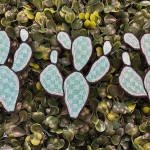 May include: Three embroidered cactus patches with a light teal and white checkered pattern, outlined in white and dark brown. The patches are arranged on a bed of green leaves.