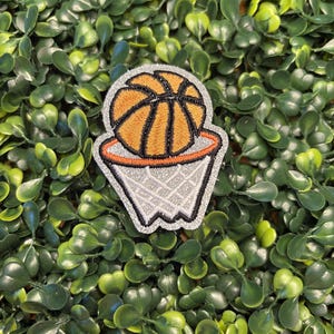 May include: A basketball patch featuring an orange basketball with black lines above a silver and black basketball hoop. The patch is set against a backdrop of green foliage.