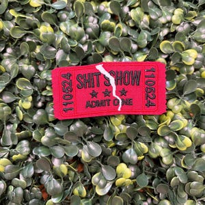 May include: A red ticket-shaped patch with the words "SHIT SHOW" and "ADMIT ONE" in black. The patch has a torn design and is set against a backdrop of green foliage.