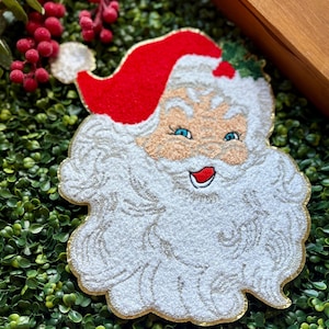 May include: A festive Santa Claus face ornament with a red hat, white beard, and blue eyes. The ornament has a gold trim and is set against a green background, perfect for holiday decorations.