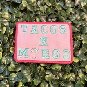 May include: A rectangular patch with the words "TACOS N MRGS" embroidered in light blue on a pink background. The patch has a red border and a margarita glass graphic. The patch is set against a backdrop of green foliage.