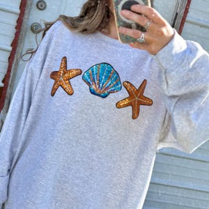May include: A light gray sweatshirt with sequined seashell and starfish designs. The sweatshirt features a blue and orange sequined seashell in the center, flanked by two orange starfish. A trucker hat with the word "CITY" is visible.