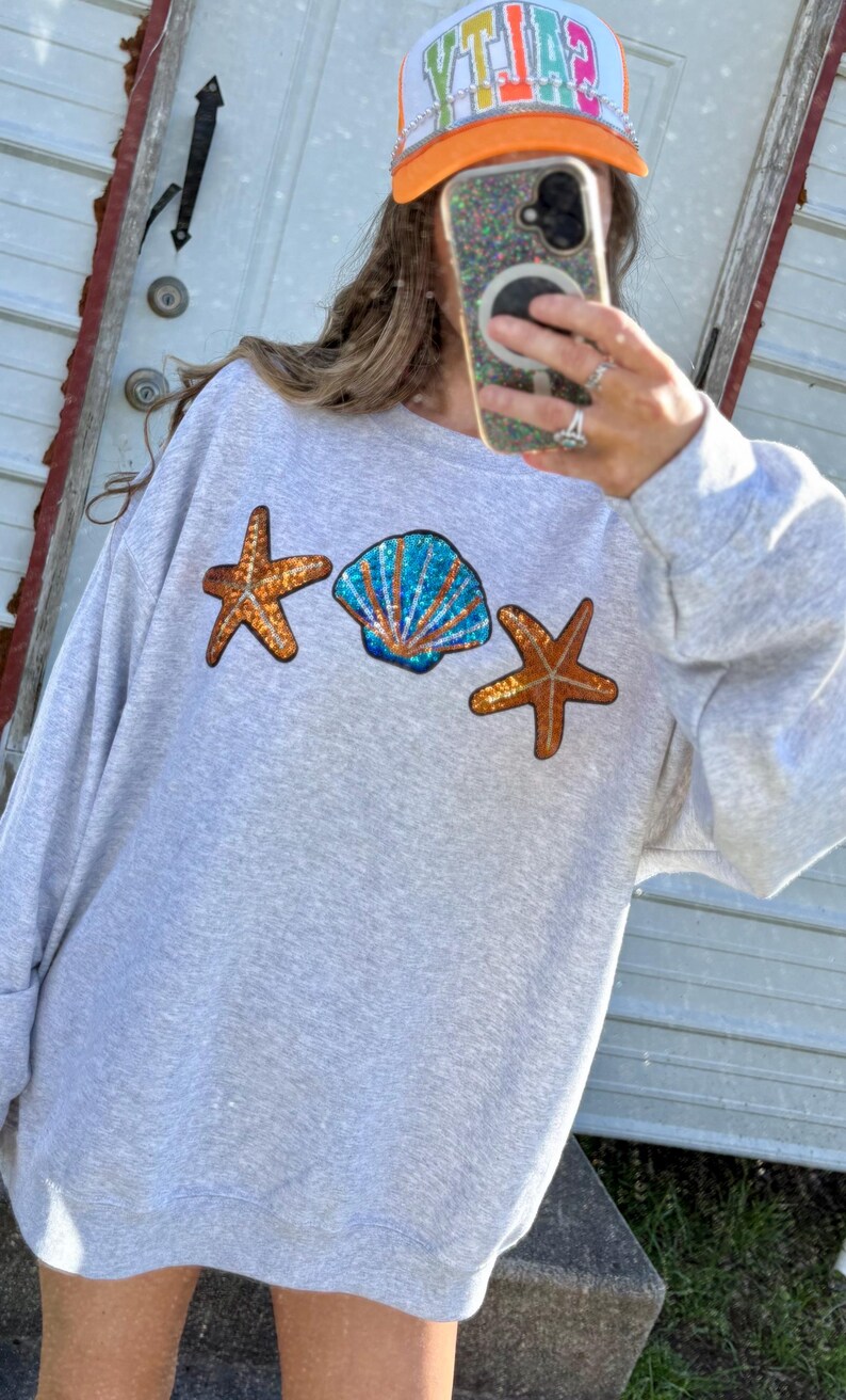 May include: A light gray sweatshirt with sequined seashell and starfish designs. The sweatshirt features a blue and orange sequined seashell in the center, flanked by two orange starfish. A trucker hat with the word "CITY" is visible.