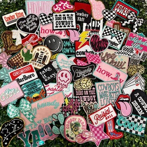 May include: A collection of embroidered patches with a country and western theme. The patches feature phrases like "Cowboy Killer," "Howdy," and "Raisin' Hell," along with cowboy boots, hats, and hearts. Colors include pink, black, white, and turquoise.