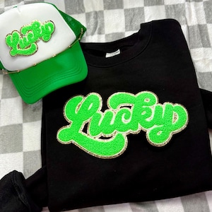 May include: A black sweatshirt and a green and white trucker hat. Both items feature the word "Lucky" in a bright green, textured font with gold trim. The hat has a green mesh back and a white front panel.