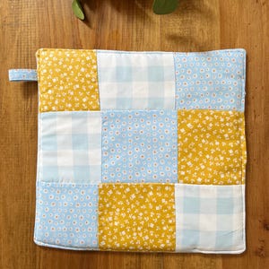 May include: A patchwork oven mitt featuring a repeating pattern of yellow floral, light blue gingham, and light blue floral fabric squares. A small fabric loop is attached to the top left corner. The mitt is edged with white fabric.