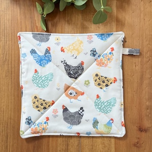 May include: Square cream-coloured pot holder featuring a repeating pattern of chickens in various colours. The chickens are in blue, yellow, black, and orange. A small loop is attached to one corner for hanging.