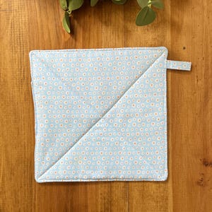 May include: A square, light blue pot holder with a white and orange floral pattern. The pot holder has a small loop for hanging and is diagonally stitched. The background is a wooden surface.
