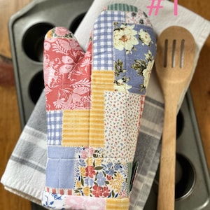 Puede incluir: Un guante de cocina de patchwork con estampados florales y de cuadros en rojo, azul, amarillo y blanco. También se ven una cuchara de madera y un molde para muffins gris. El guante está sobre una toalla a rayas blancas y grises.