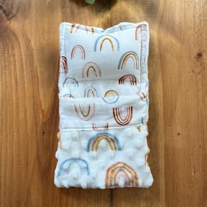 May include: A white, plush baby item with a rainbow pattern in shades of blue, orange, and brown. The item is rectangular and folded, with a textured, dimpled fabric on the bottom layer. The rainbow design is repeated across the item.