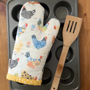 May include: A quilted oven mitt featuring a repeating pattern of colorful chickens in shades of blue, yellow, black, and orange on a white background, finished with a yellow floral trim, paired with a light brown wooden slotted spatula.