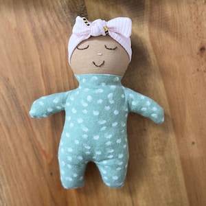 Könnte beinhalten: Handgemachte Puppe mit hellbraunem Gesicht, geschlossenen Augen und einem aufgestickten Lächeln. Sie trägt einen hellgrünen Strampler mit weißen Punkten und ein rosa Stirnband mit Schleife. Die Puppe ist aus weichem Stoff.