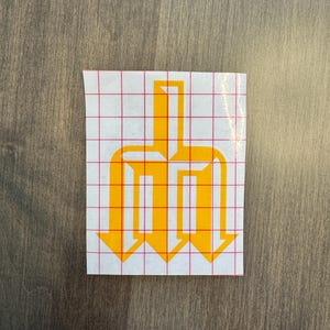 May include: A yellow vinyl decal featuring a stylized trident or pitchfork symbol with sharp, angular lines. The design is printed on a white backing sheet overlaid with a red grid pattern, commonly used for precise alignment during application.