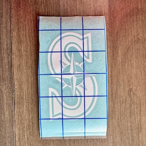 May include: A white vinyl decal featuring a stylized letter S with a compass rose emblem integrated into the center. The decal is applied to a light blue backing paper with a blue grid pattern overlay, designed for precise alignment and application.