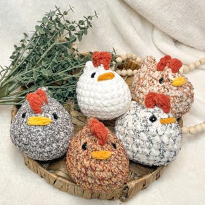 May include: Five handmade crochet chickens in a woven basket. The chickens are various colors, including white, gray, and brown, with orange combs and yellow beaks. The basket is on a white surface with greenery and wooden beads.