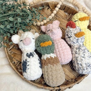 May include: A collection of handmade crochet farm animal toys, including a cow, duck, pig, chicken, and duckling. The toys are white, pink, green, yellow, and black, with orange beaks and accents. They are arranged in a woven basket.