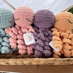 May include: Four handmade crochet octopus plush toys in a woven basket. The plushies are in shades of blue, pink, purple, and orange, each with closed-eye details. Each octopus has multiple curled tentacles. The basket has a woven handle.