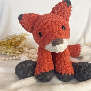 May include: A handmade, plush fox toy in shades of orange, white, and gray. The fox has a fluffy texture, black eyes, and a white-tipped tail. The fox's ears and feet are accented with gray. The toy is sitting on a white surface.