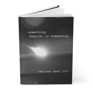 May include: A hardcover journal with a black and white cover. The cover features the text "something magical is happening" and "can you feel it?" in a simple, sans-serif font. The journal's pages are white.
