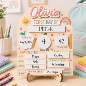 May include: A wooden arch-shaped sign for the first day of school featuring the name Olivia in pink letters. The board includes sections for date, age, height, favourites, and teacher, with colourful graphics of a sun, rainbow, apple, and pencil.