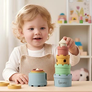 May include: A child plays with a colorful stacking toy. The toy includes various pastel-colored cups and rings, forming a tower. The base of the tower has the name "lucas" printed on it. The toy is on a light wooden table.