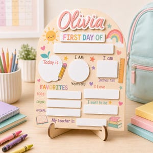 Peut inclure: Un panneau en bois clair en forme d'arche avec le prénom Olivia en haut. Il comporte des sections pour First Day Of, Today is, I Am, Years Old, I am, Inches tall, Favorites, Food, Color, Animal, I Love, I want to be et My teacher is.