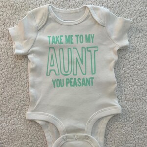 May include: A white short-sleeved baby bodysuit featuring the text "TAKE ME TO MY AUNT YOU PEASANT" printed in a light mint green color on the chest. The garment has a round neckline and three snap closures at the bottom for easy access.