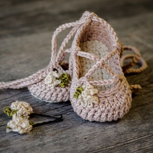 Pode incluir: Um par de sapatos de bebé de crochet rosa com enfeites de flores brancas e verdes. Os sapatos têm atacadores que se amarram à volta dos tornozelos.