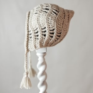 Peut inclure: Un bonnet de bébé en crochet avec un long pompon noué. Le bonnet est fait de fil de couleur crème et a un design simple à points ouverts.