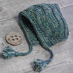 May include: A green knitted baby bonnet with a cable knit pattern and two long tassels. The bonnet is lying on a wooden surface with a wooden button with the text "Two Girls Patterns" on it.