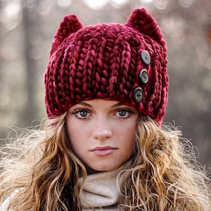 May include: A red knitted beanie with cat ears and three black buttons. The beanie is made from a thick, chunky yarn.