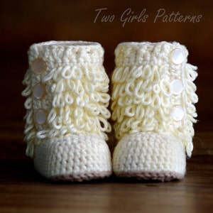 May include: A pair of cream-colored, crocheted baby booties. The booties have a textured, looped design around the ankle and are secured with three pearlescent buttons. The soles are a contrasting light brown color. The text "Two Girls Patterns" is in the upper right corner.