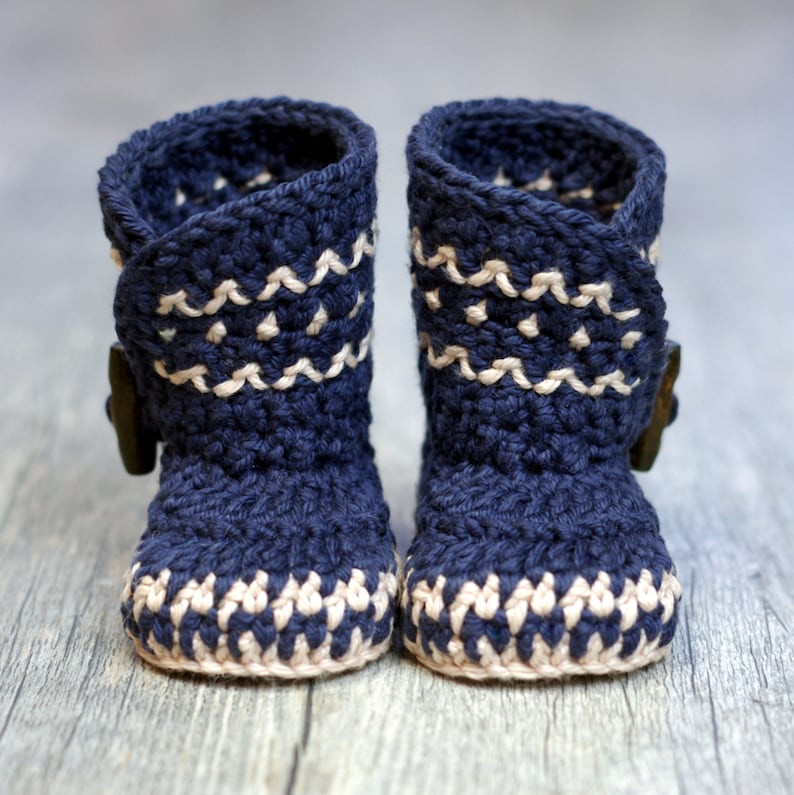 K&ouml;nnte beinhalten: Ein Paar marineblaue geh&auml;kelte Babyschuhe mit wei&szlig;en Akzenten. Die Schuhe haben einen umgeschlagenen Bund und ein Knopfdetail.