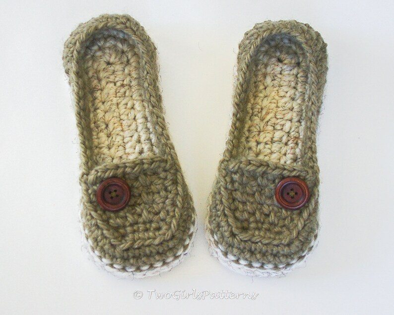May include: A pair of crocheted slippers in a green and beige color scheme. The slippers have a button detail on the front and a white trim around the edge.