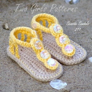 Pu&ograve; includere: Un paio di sandali per neonati lavorati all'uncinetto in giallo chiaro e beige. I sandali presentano un design a cinturino a T decorato con tre gemme in plastica trasparente sfaccettate e iridescenti. Testo: Two Girls Patterns Seaside Sandals 211.