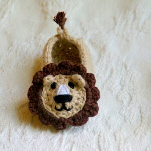 May include: A pair of crocheted baby booties with a lion face design. The booties are beige with brown lion faces and a brown mane.