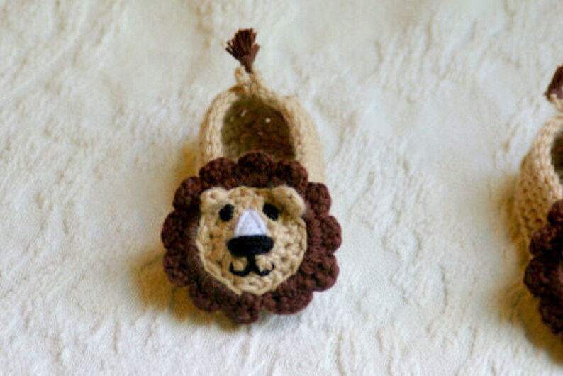 May include: A pair of crocheted baby booties with a lion face design. The booties are beige with brown lion faces and a brown mane.