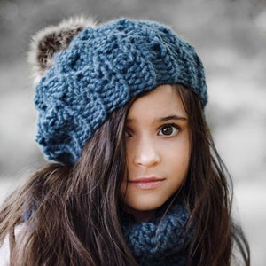Pode incluir: Um gorro azul de malha com um pompom felpudo no topo. O gorro é usado por uma pessoa jovem com cabelo castanho comprido.